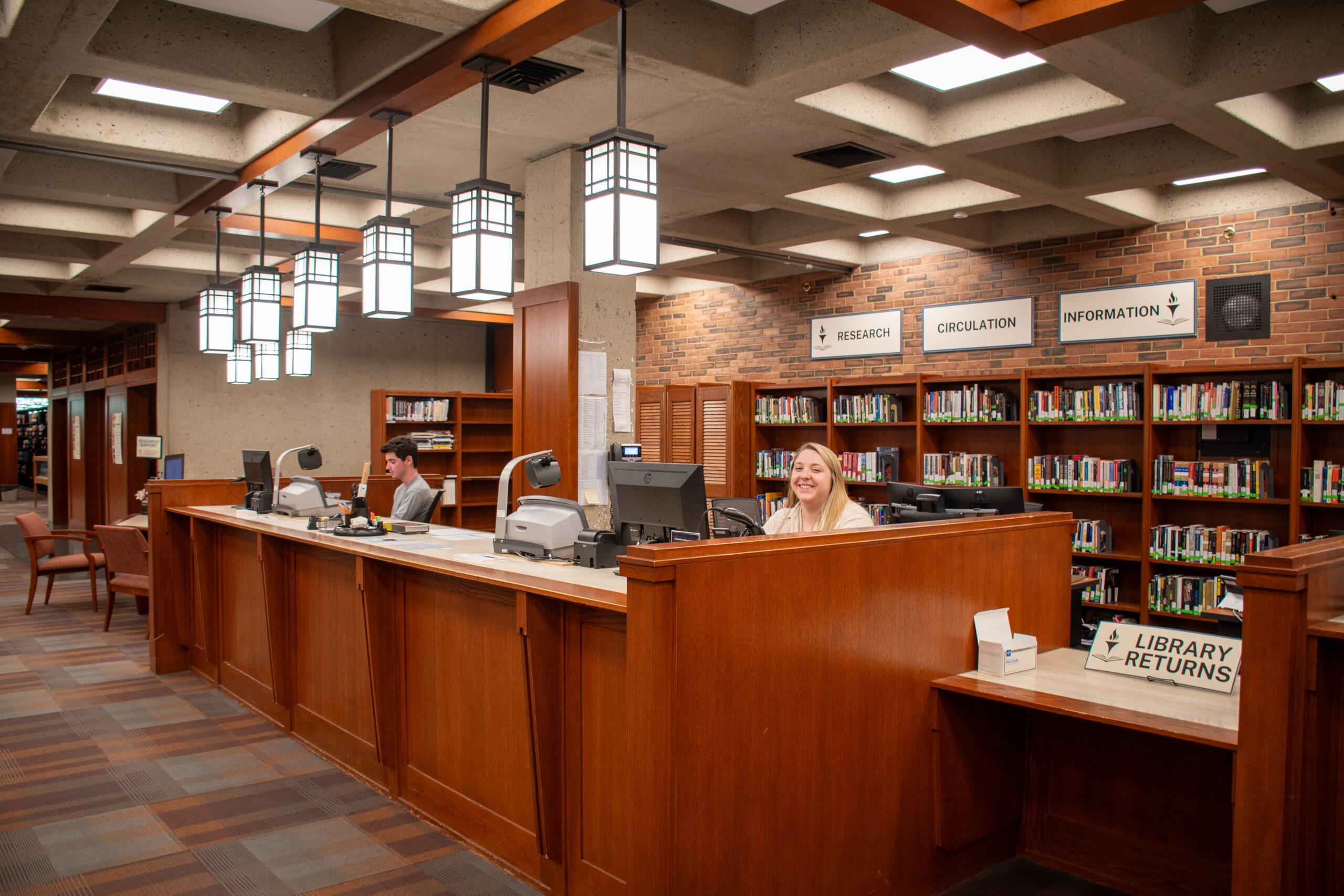 Two staff people working at the library circulation desk, with one smiling towards the viewer. There are signs for Library Returns, Research, Circulation, and Information. The circulation desk contains computers, scanners, and other office tools. Behind the staff people are shelves of books.