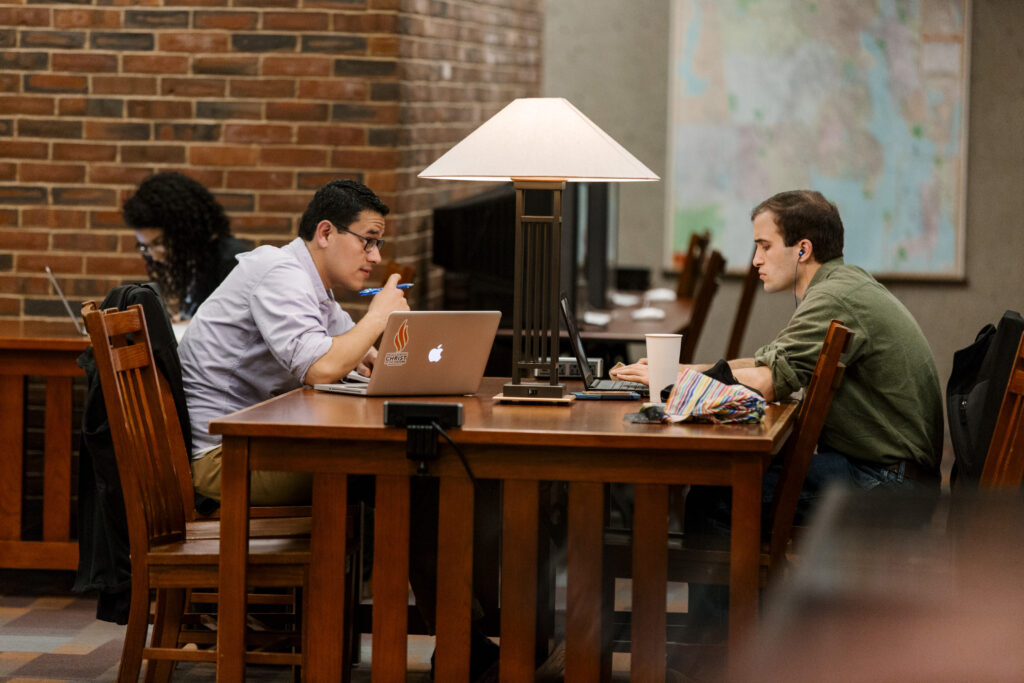 Two students study on laptops at a table in the library.