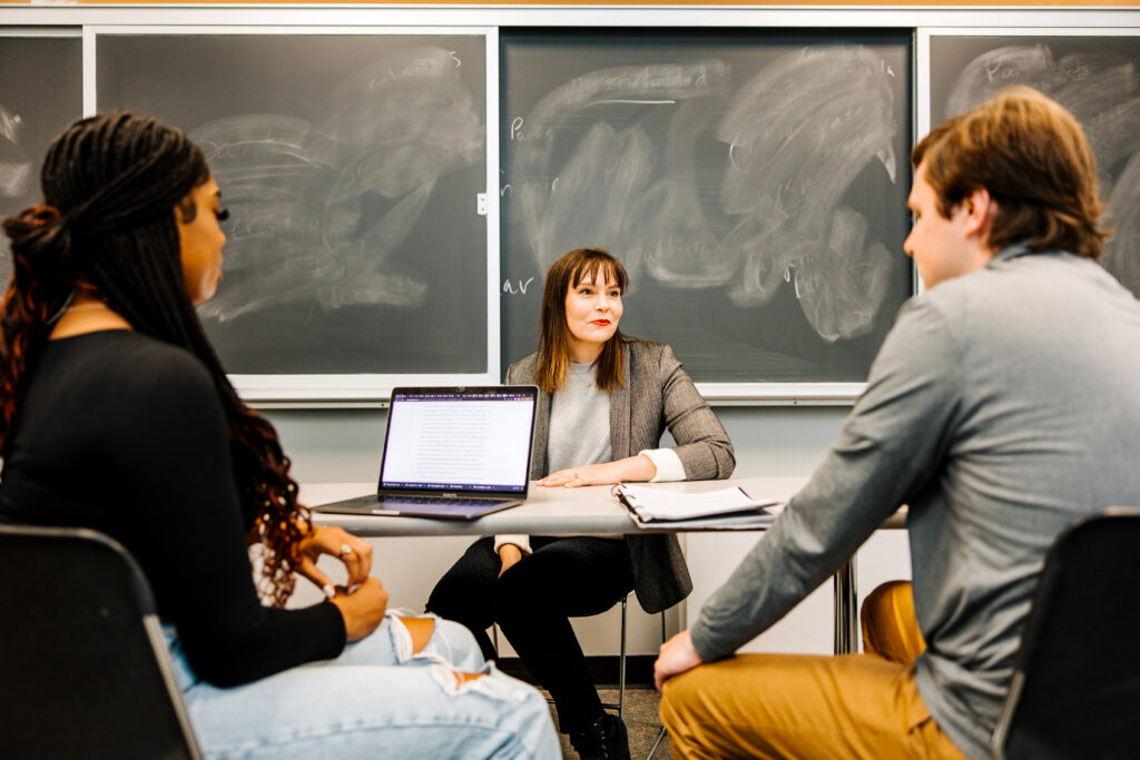 Two students and a professor have a discussion together in a classroom.