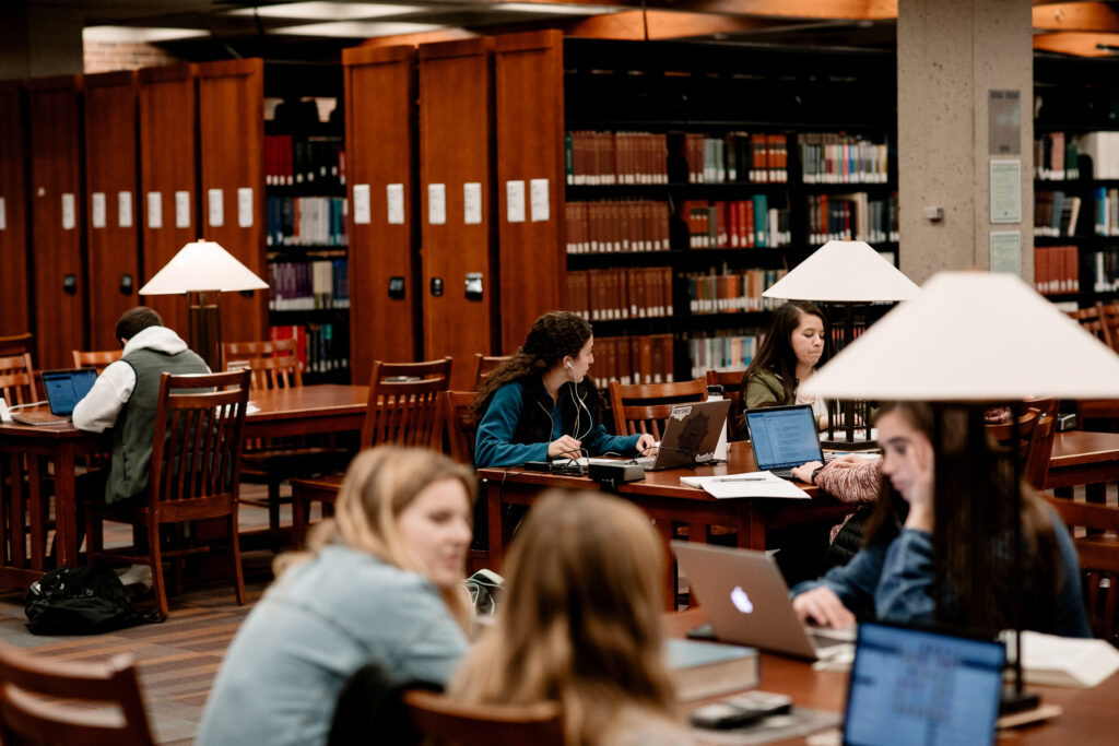 Students in groups across multiples tables sit and work in the library main level floor.