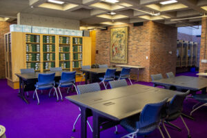 The Fogarty Reading room containing modular tables, blue ergonomic chairs, and archival bookshelves, complemented by a large tapestry on a brick wall. The room has a unique purple carpet.