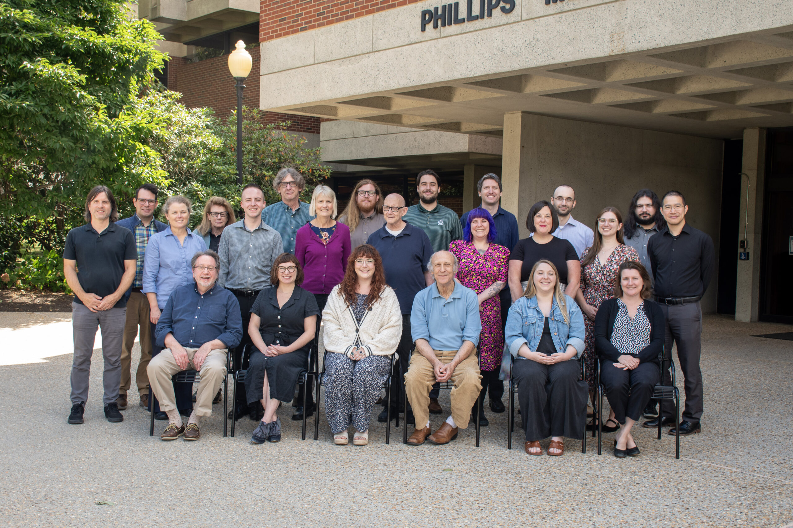 The Phillips Memorial Library staff grouped together, smiling towards the viewer. They are in front of the Phillips Memorial Library on a sunny day.