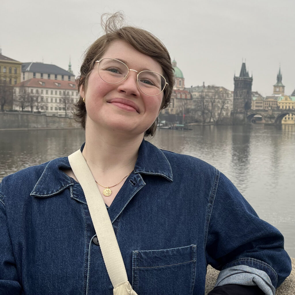 Photo of Rebecca Doctor, an archivist, standing in front of a river with a European looking city behind her. She has short brown hair and glasses.