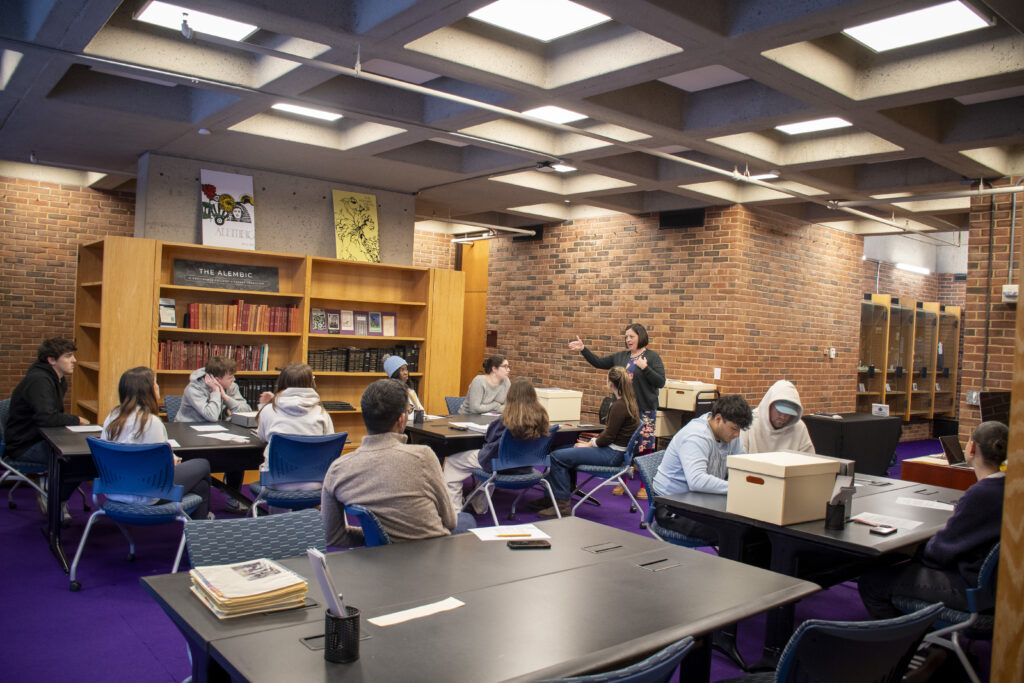 A group of students sits at tables in a Fogarty Reading Room, listening to a woman standing and speaking at the front. The space has brick walls, wooden bookshelves filled with bound volumes, and posters displayed above the shelves. Several students are examining archival boxes and documents, while others face the instructor. The room has a purple carpet and a grid-patterned ceiling with recessed lighting.