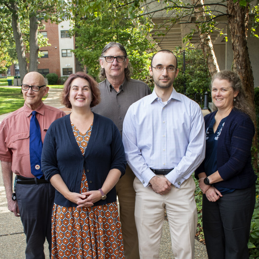 Five adults stand together outside on a tree-lined campus path, posing for a group photo. The group includes three men and two women, all dressed in business casual attire. A brick building and people seated at tables are visible in the background.