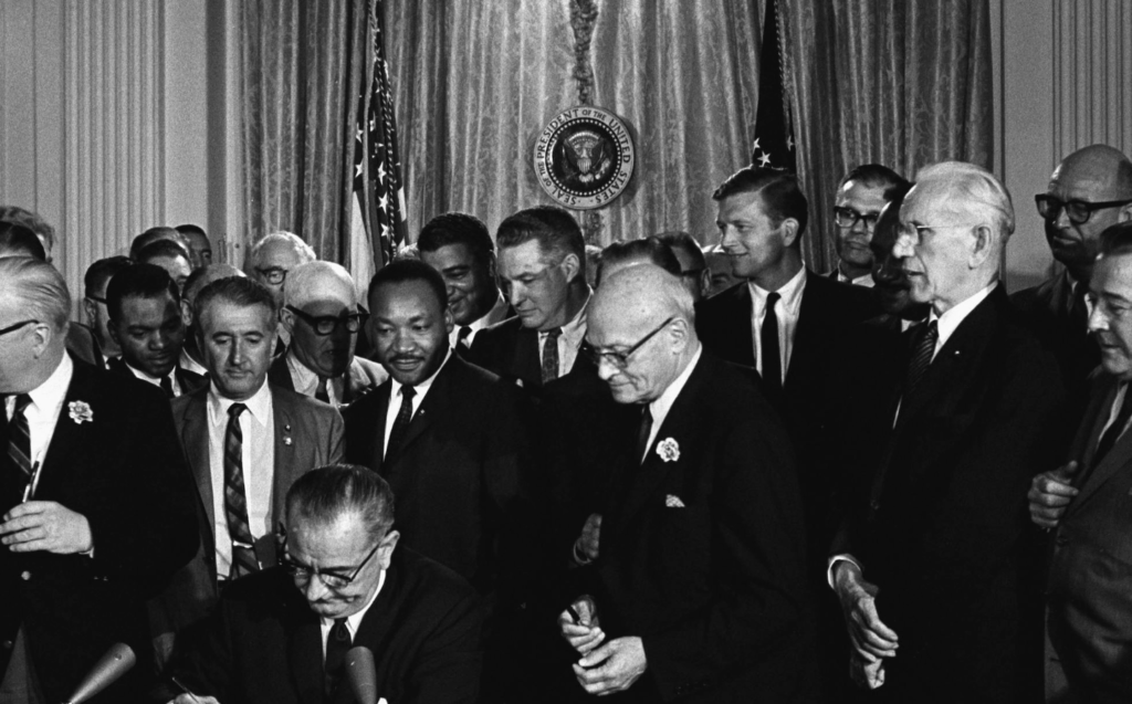 A black-and-white photograph of President Lyndon B. Johnson seated at a desk signing the Voting Rights Act of 1965. He is surrounded by a large group of men, including civil rights leaders and politicians. Martin Luther King Jr. stands near the center, watching Johnson sign the document. The presidential seal and U.S. flags are visible in the background.