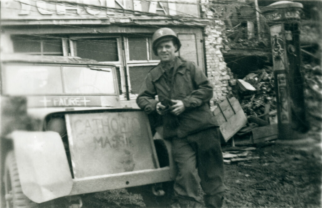 Fr. Doyle leans against a Jeep car, which has the words “padre” and “Catholic Mass” written on it 
