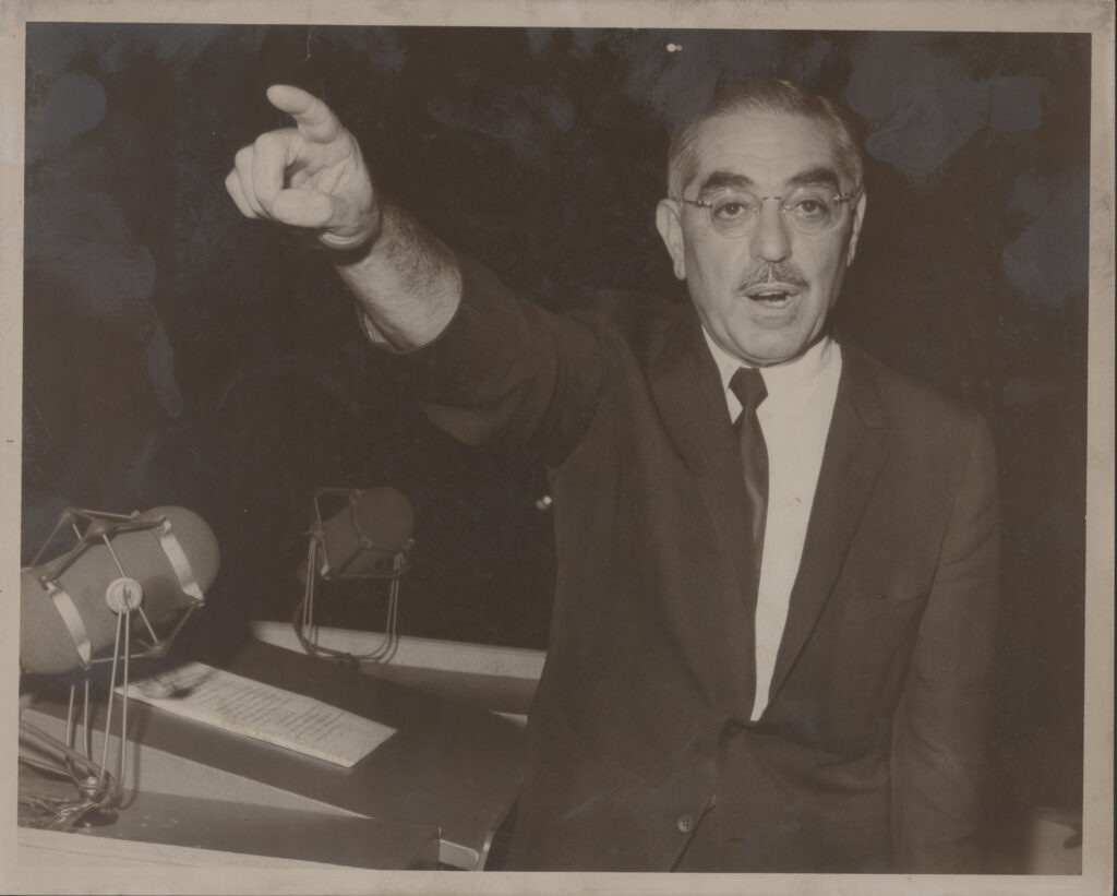 Black and white photograph of John O. Pastore in a suit and tie, standing behind a desk with microphones and papers. He is pointing forward with an outstretched arm and looking directly at the camera.