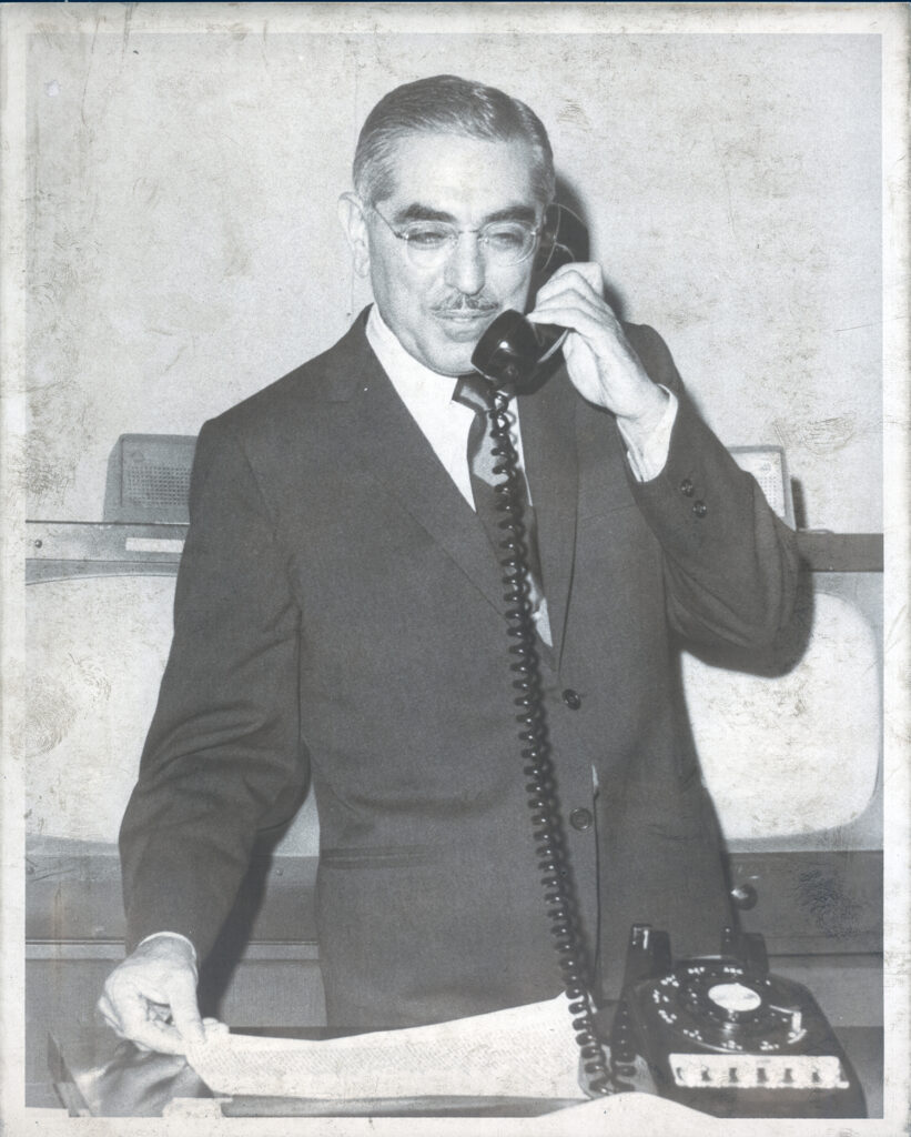 Black and white photograph of John O. Pastore in a suit and tie, standing behind a desk while holding a rotary dial telephone receiver to his ear. He is looking down at papers held in his other hand.