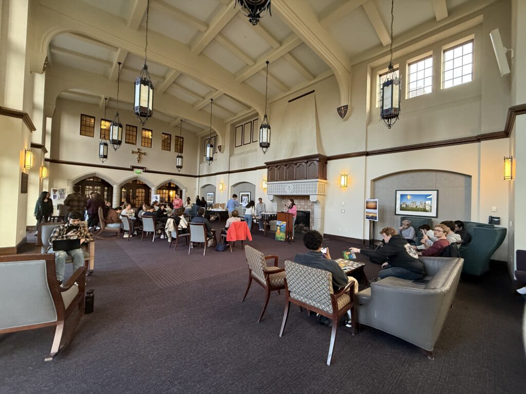 The Ruane Great Room set up for The Alembic 2026 launch event with around 20 students sitting facing a podium with a female student in pink speaking at the microphone.