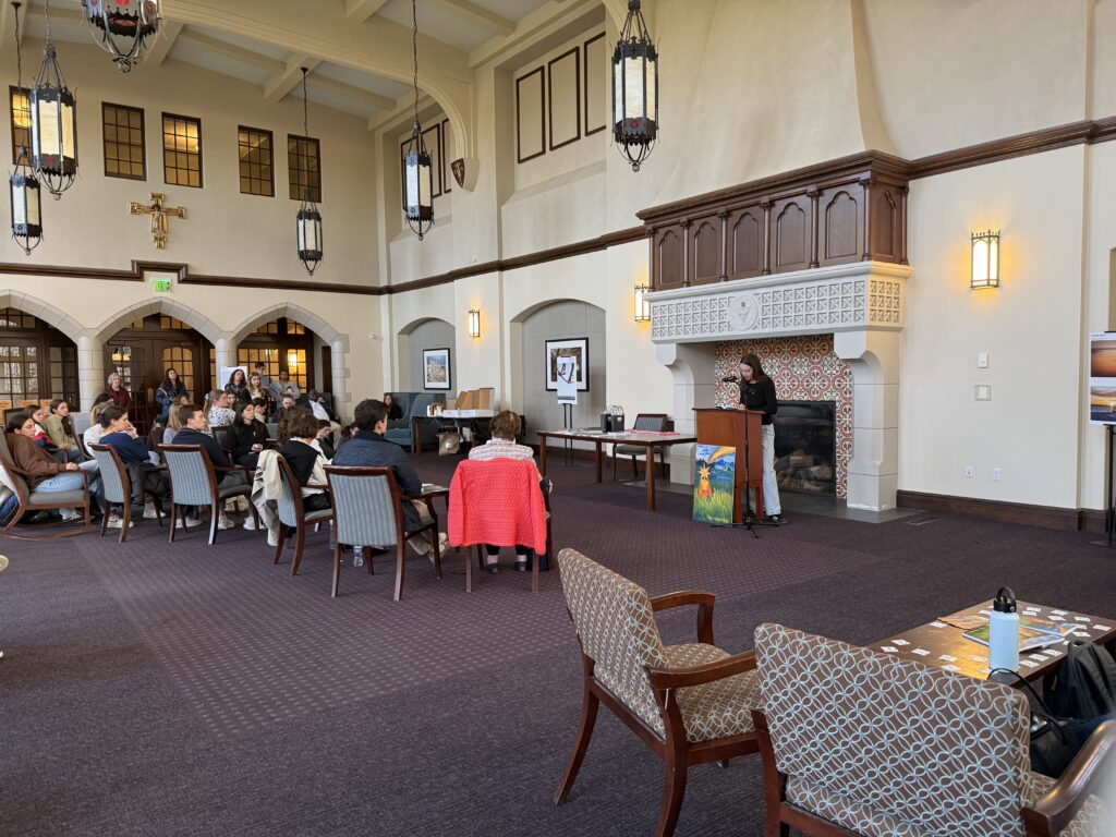 The Ruane Great Room set up for The Alembic 2026 launch event with around 20 students sitting and standing facing a podium with a female student in a black long sleeve reading a work from The Alembic.