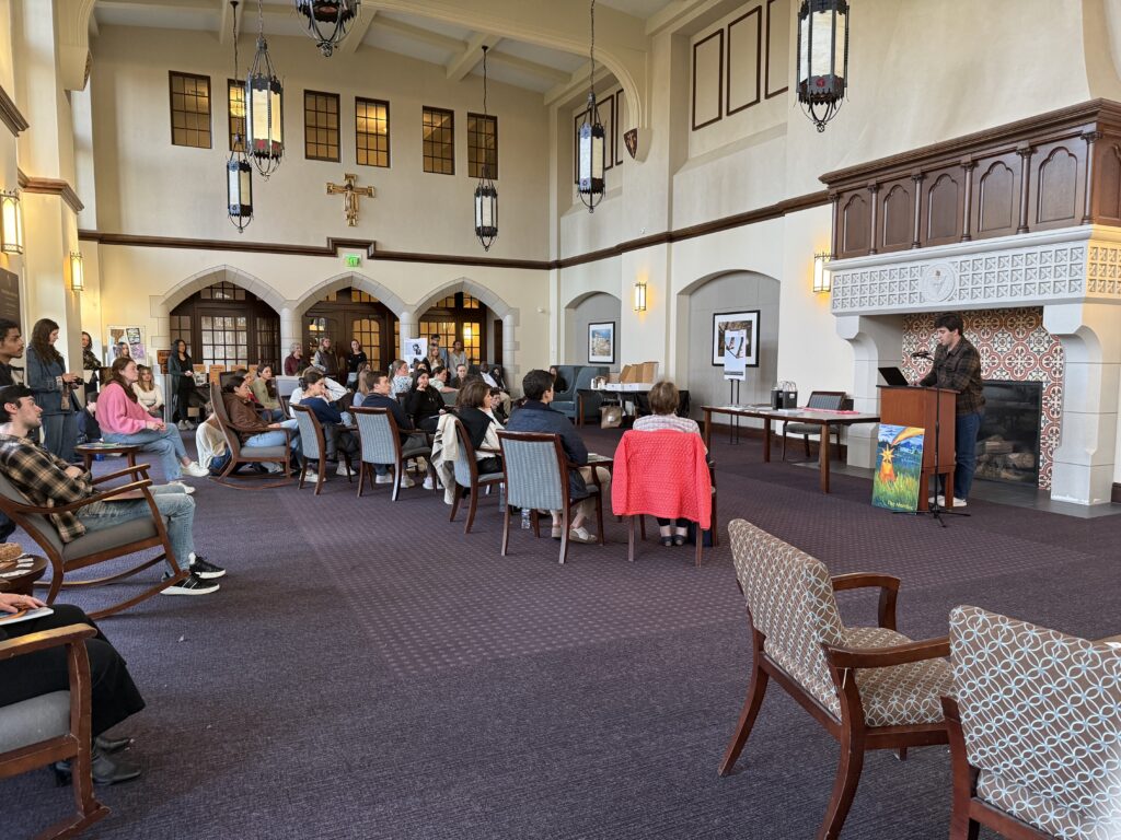 The Ruane Great Room set up for The Alembic 2026 launch event with around 20 students sitting and standing facing a podium with a male student in dark flannel reading a work from The Alembic.