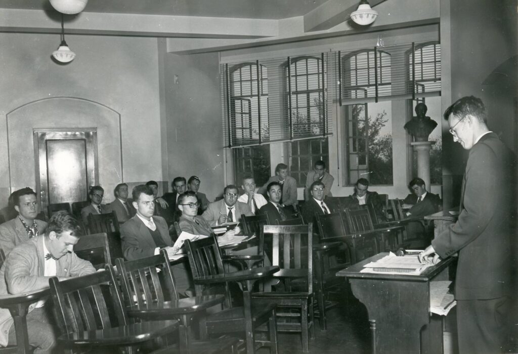 Student stands at podium, presenting to a group of students in a classroom setting.