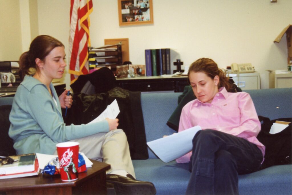 Two students sit on a couch and review papers in an office.