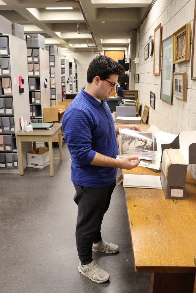 Student stands in a collections storage room, flipping through documents in folders next to a table with boxes.