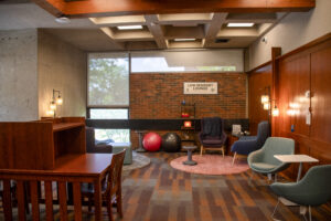 A low sensory lounge in a library with warm wood paneling, brick walls, soft lighting, study carrels, armchairs, small tables, rugs, exercise balls, and calming sensory items. A sign on the back wall reads “Low Sensory Lounge.”