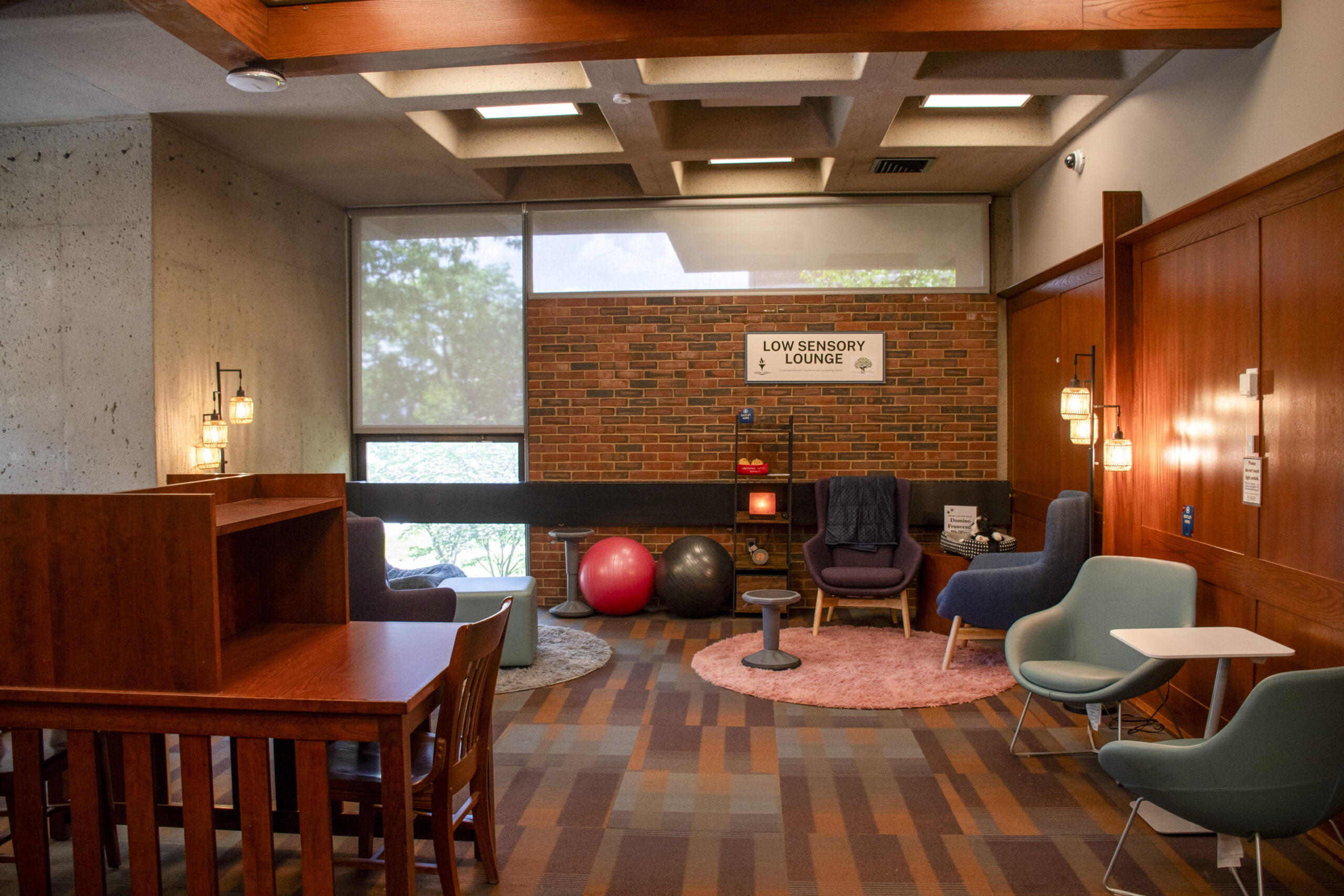 A low sensory lounge in a library with warm wood paneling, brick walls, soft lighting, study carrels, armchairs, small tables, rugs, exercise balls, and calming sensory items. A sign on the back wall reads “Low Sensory Lounge.”