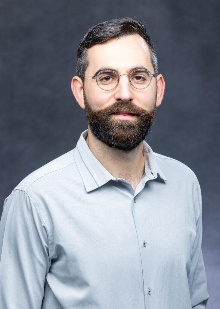A professional headshot of Dr. Thomas H Ptashnik wearing glasses and a light blue button-down shirt against a dark gray studio backdrop.