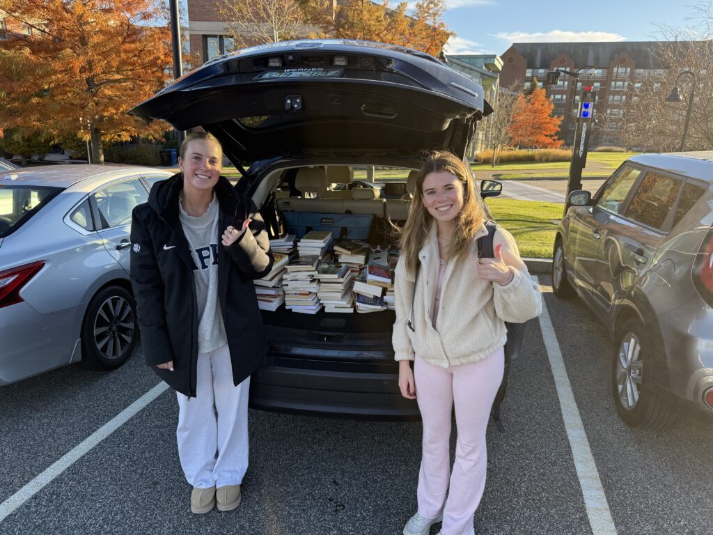 Araceli Cloutier and Katherine Greek stand beside an open car trunk filled with donated books on the Providence College campus.