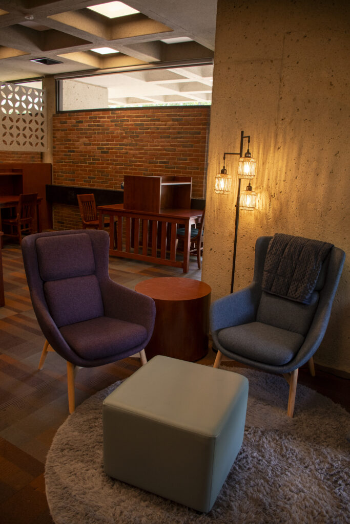 Low-sensory lounge area featuring two upholstered lounge chairs, a cushioned ottoman, and a small round table arranged on a soft circular rug. Warm pendant lighting and natural wood furnishings create a calming atmosphere, with quiet study carrels and brick walls visible in the background.