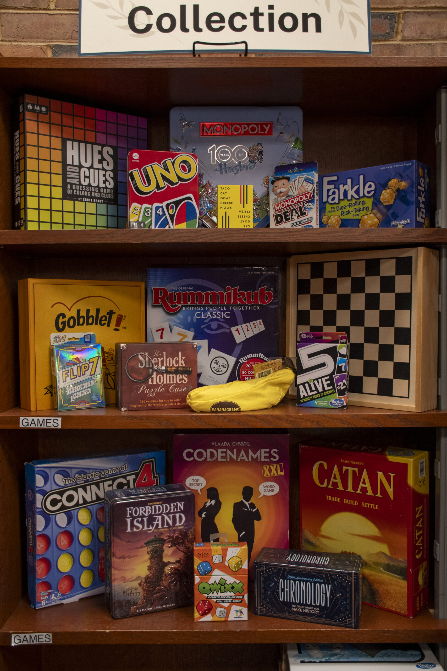 Wooden library shelving displaying a variety of games available in the Wellness Collection. Visible titles include Hues and Cues, UNO, Monopoly Deal, Farkle, Gobblet, Rummikub, Connect 4, Codenames XXL, Catan, Forbidden Island, Qwixx, and Chronology, along with card games, puzzles, and a checkerboard. A sign above the shelf reads “Collection.”