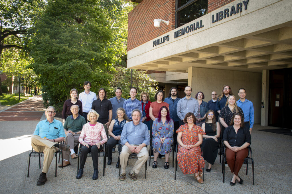 The Phillips Memorial Library staff grouped together, smiling towards the viewer. They are in front of the Phillips Memorial Library on a sunny day.